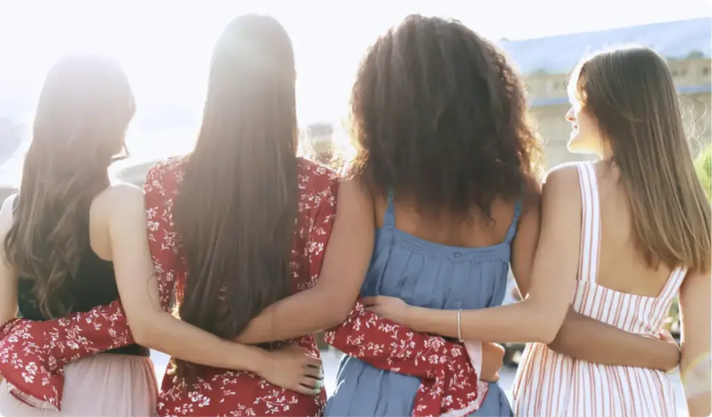Four women stand side by side with their arms around each other, facing the sunlight, symbolizing connection and support in recovery.