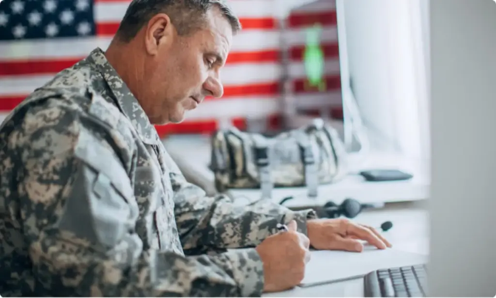 A military veteran in uniform sits at a desk writing, with an American flag in the background.